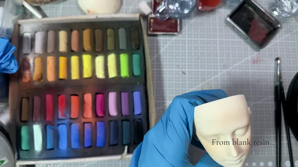 A blue-gloved hand supports a light, mostly unpainted BJD head beside a color palette.