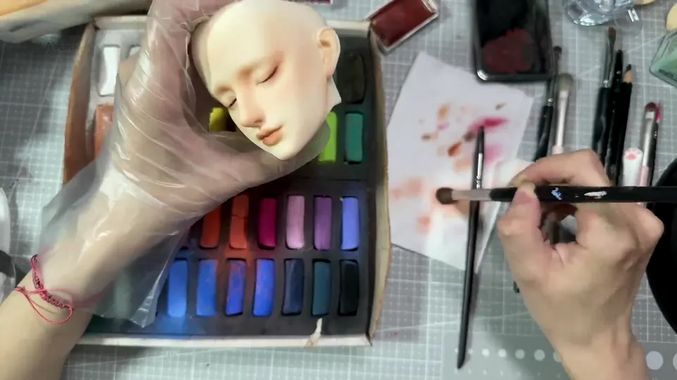 A BJD head with soft pink shading is held beside a soft brush and paper bearing color marks.