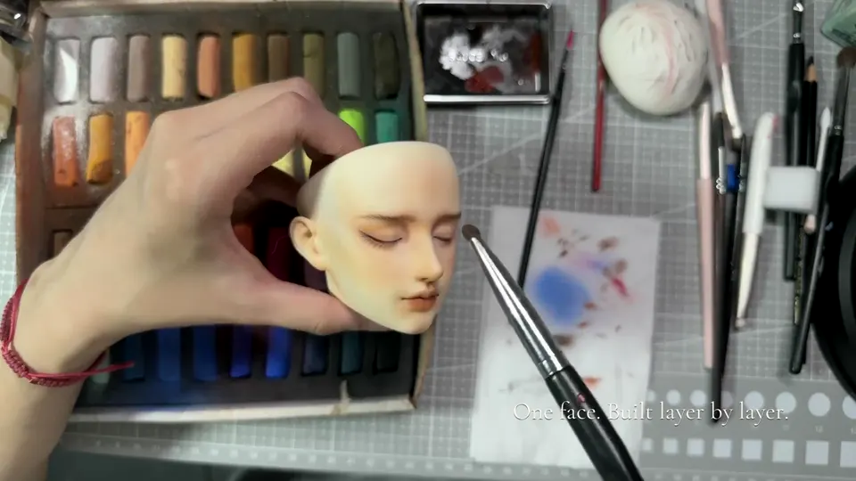 A hand holds a BJD head with defined brows, lash lines and lips while a brush is positioned near the outer eye.