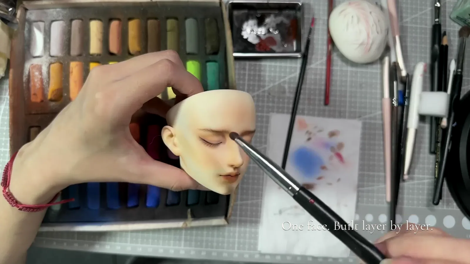 A hand holds a BJD head while a brush works near the eye, with color sticks and tools on the table.